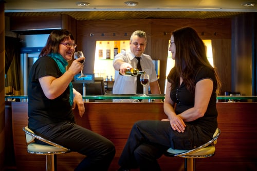 To damer som koser seg med en drink i baren, bartender som fyller opp et glassvin.