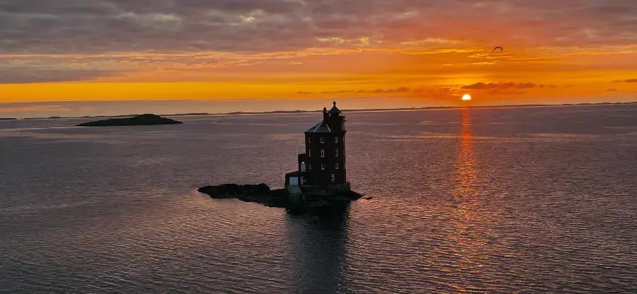 The image depicts a serene sunset over the ocean with a lighthouse on the right, a small island in the middle, and bright clouds in the sky.