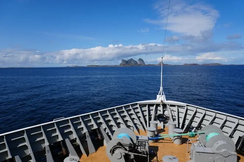 a view from the deck of a ship looking out at the sea. There are large rocks visible in the distance on the horizon under a blue sky with some scattered clouds. The deck has safety railings, and there are various pieces of equipment and machinery visible.