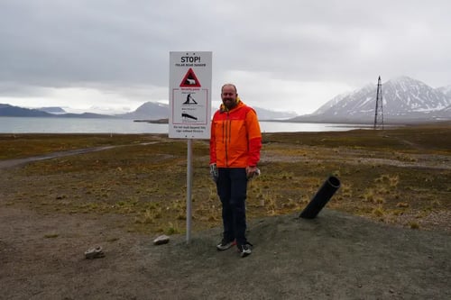 an individual standing next to a signpost in a barren landscape. The person is wearing an orange jacket, dark pants, and dark shoes, and is facing the camera. The signpost has a white background with a red border and the word 'STOP!' at the top in bold black letters