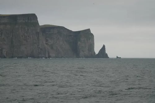 a coastal scene with a large cliff on the left and a smaller rock formation on the right. The sea is visible in the foreground with waves gently lapping at the shore. The sky is overcast, contributing to the moody atmosphere of the scene.
