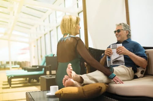 a man and a woman sitting on a sofa in a modern living space with a high ceiling and glass windows. The man is wearing a blue shirt and shorts, holding a book or magazine, while the woman, with her back to the camera, is wearing a sparkling dress.