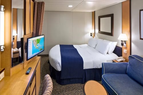 a neatly arranged hotel room. There is a large bed with blue bedding and two white pillows. A flat-screen TV is mounted on the wall, displaying a map. A sofa is positioned next to the bed, and there is a small table with a lamp and a remote control on it.