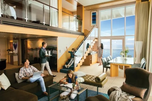 modern living room with a large window offering a view of the ocean. There's a staircase with glass railings leading to an upper level. In the foreground, there's a seating area with a couch, armchairs, and a coffee table.