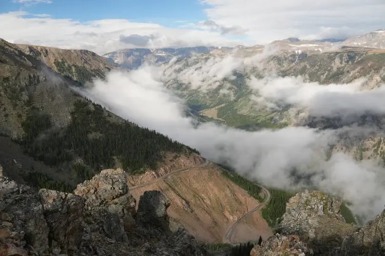 a breathtaking view of a mountainous landscape. A winding road cuts through the terrain, disappearing into a blanket of clouds that hover between the mountains. The mountains themselves are a mix of greenery and rocky outcrops, hinting at the ruggedness of the terrain.
