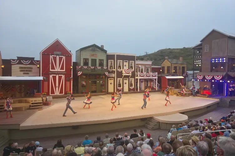 A lively scene of a stage performance in a rural setting, with actors dressed in colorful costumes, and an audience watching the show.