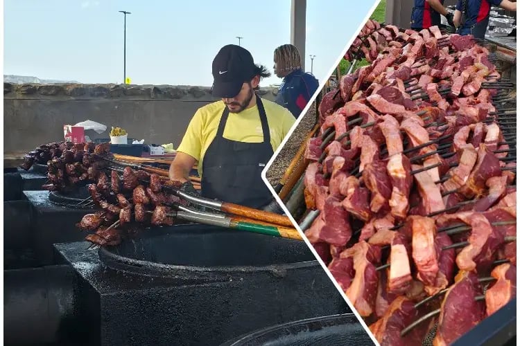 a person wearing a black cap, a yellow shirt, and a black apron, working behind a grill with skewers of meat. The skewers are arranged on a rack, and the grill appears to be outdoors. The background suggests a sunny day with a clear sky and a hint of buildings or structures in the distance.