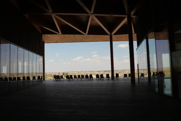 n interior space with a modern design, featuring a series of dark-colored benches arranged in rows. The space is illuminated by natural light coming through large windows that provide a view of a distant landscape.