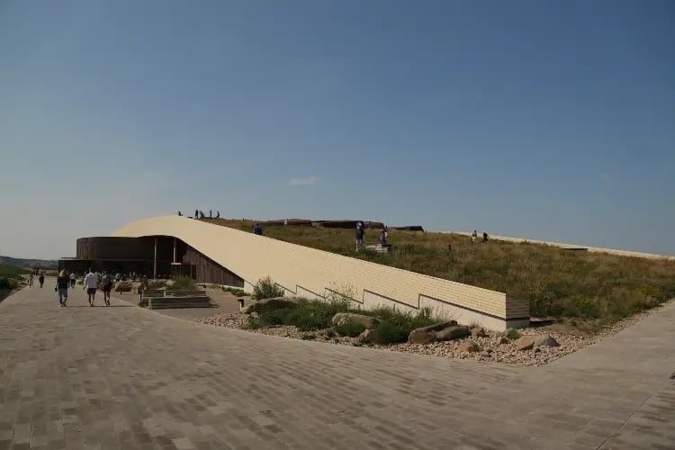 a modern building with a sloped roof and large glass windows, situated on a hill. There is a pathway leading up to the building, and a few people can be seen walking along it. The sky is clear and blue, suggesting fair weather.