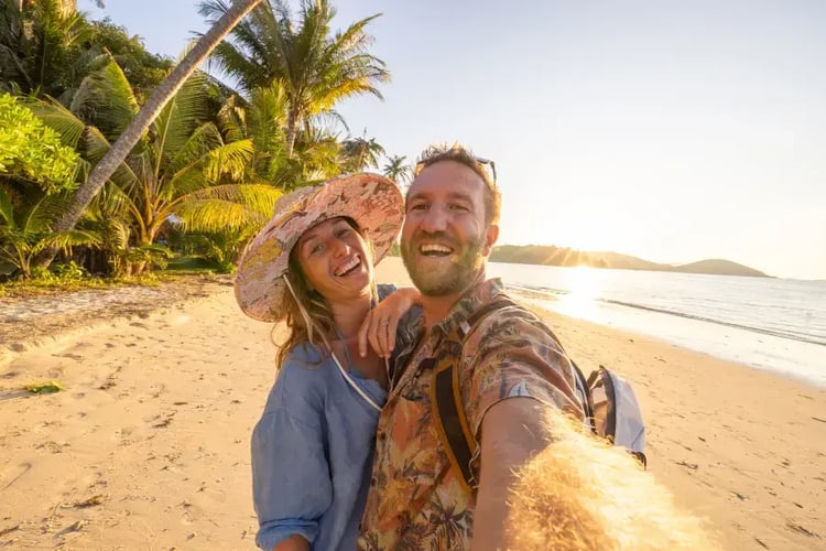 two people taking a selfie on a sandy beach during sunset. The person on the left is wearing a blue shirt and a wide-brimmed hat, while the person on the right is wearing a patterned shirt. There is a palm tree and lush greenery in the background