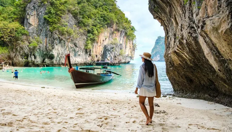 hows a serene beach scene with a person walking on the sand. In the background, there is a boat moored to a rocky cliff. The water is calm and clear, revealing the sandy bottom. The sky is partly cloudy, and there are other people visible in the distance on the beach.