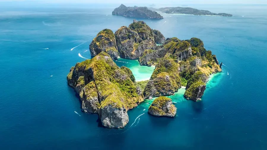 stunning aerial view of a landmass surrounded by deep blue sea water. The landmass features sharply defined, rocky formations with patches of green vegetation. There is a beautifully turquoise lake or lagoon at the center of the landmass, indicative of a volcanic origin, possibly a crater lake