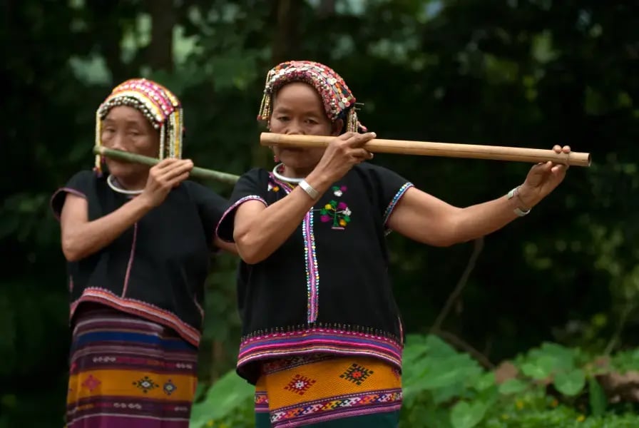 The image shows two individuals from behind, likely women, playing a traditional wooden flute. They are wearing black tops with colorful embroidery and patterned skirts. The setting appears to be outdoors with green foliage in the background.