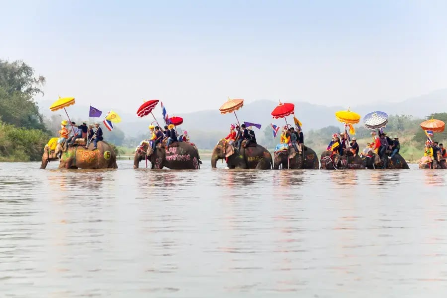 a group of people riding on the backs of elephants, each elephant carrying a person with them. The riders are adorned with colorful, oversized umbrellas, and some are holding flags. They are moving through a body of water, possibly a river.