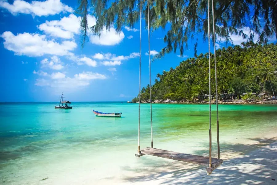 The image depicts a tropical beach scene with clear turquoise waters, white sand, and a swing suspended between two trees. In the distance, there are two boats on the water, and the sky is partly cloudy. The overall atmosphere is serene and inviting.