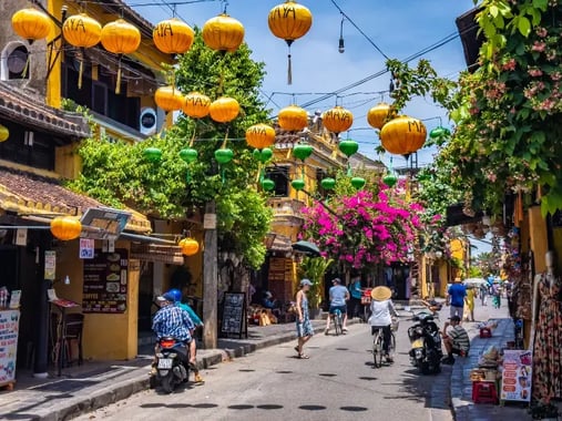 a vibrant street scene in a bustling market area. There are yellow lanterns hanging from wires above the street, creating a warm and inviting atmosphere. The street is lined with multiple small shops and restaurants, some of which have outdoor seating.