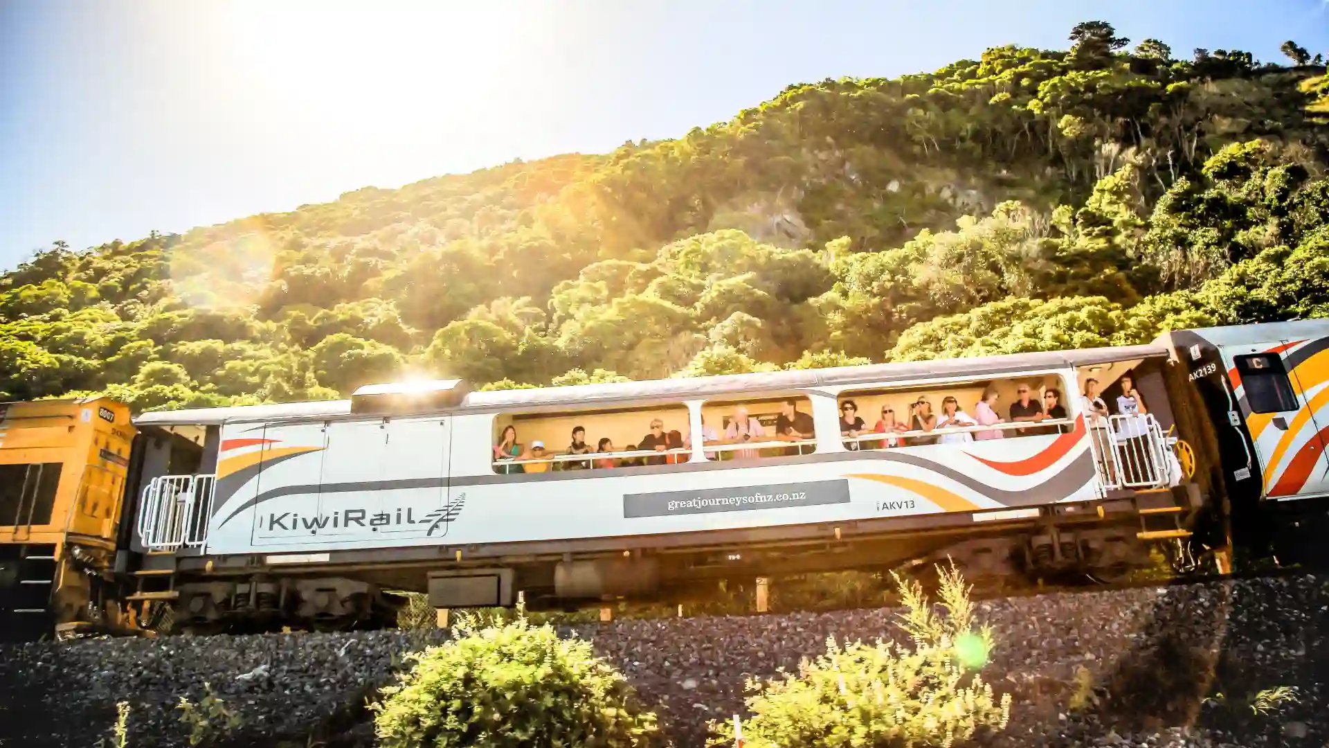 KiwiRail train with the name 'L&LX' displayed on it, traveling past a lush green hill with people on board, under a bright sun.