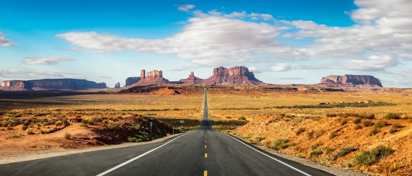 a long, straight road extending into the distance with a desert landscape on either side. The sky is clear with a few clouds, and there are large, flat-topped rock formations in the background.