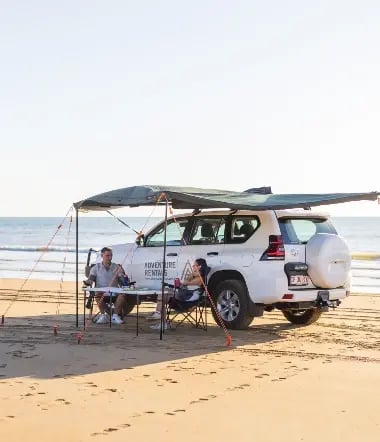 a white SUV parked on a sandy beach with a green canopy attached to the roof, providing shade. There are two individuals seated at a folding table under the canopy, with fishing rods in front of them, suggesting they are fishing.