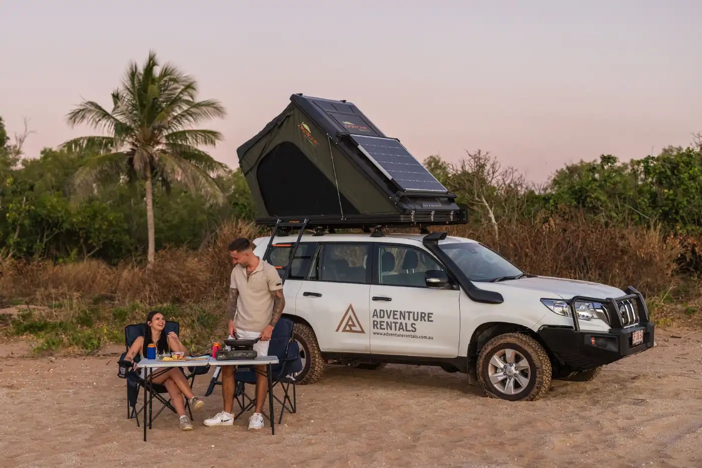 Par lager mat ved Toyota Prado med taktelt og solcellepanel på stranden ved solnedgang
