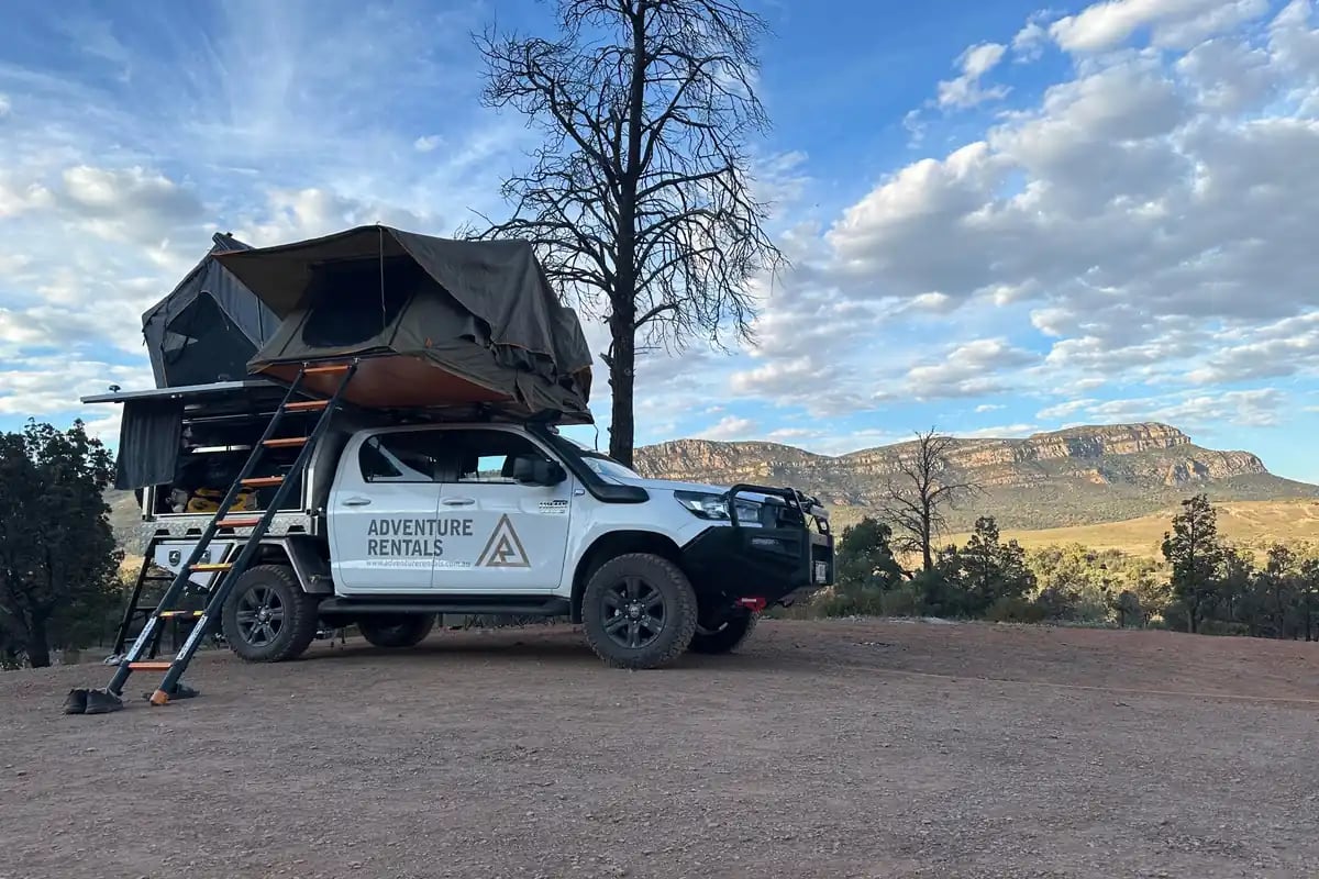off-road vehicle with a cargo area on top, equipped with a pop-up tent and ladders for camping. The vehicle is branded with 'ADVENTURE RENTALS', parked on a dirt road with a clear view of a mountainous landscape and a few trees in the background.