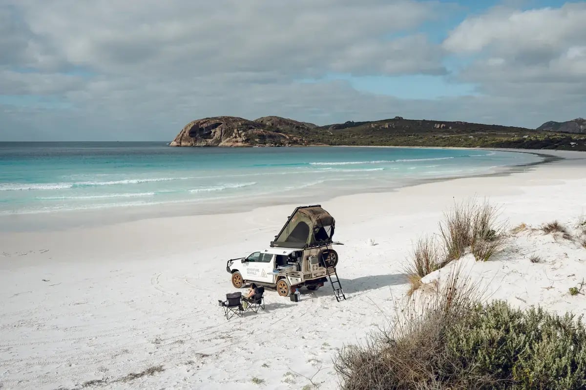 a white off-road vehicle with a tent attached, parked on a sandy beach with clear blue waters and turquoise shallows. A rocky island is visible in the distance under a partly cloudy sky.
