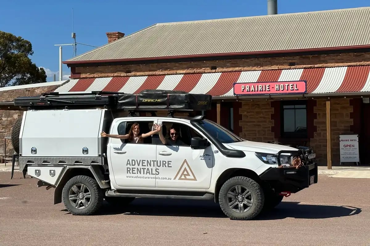 a white off-road vehicle with 'ADVENTURE RENTALS' written on the side, parked in front of a building with a red and white striped awning that reads 'PRAIRIE HOTEL'. There is also a smaller sign that appears to say 'OPEN BRO'. Two people are standing behind the vehicle, waving.