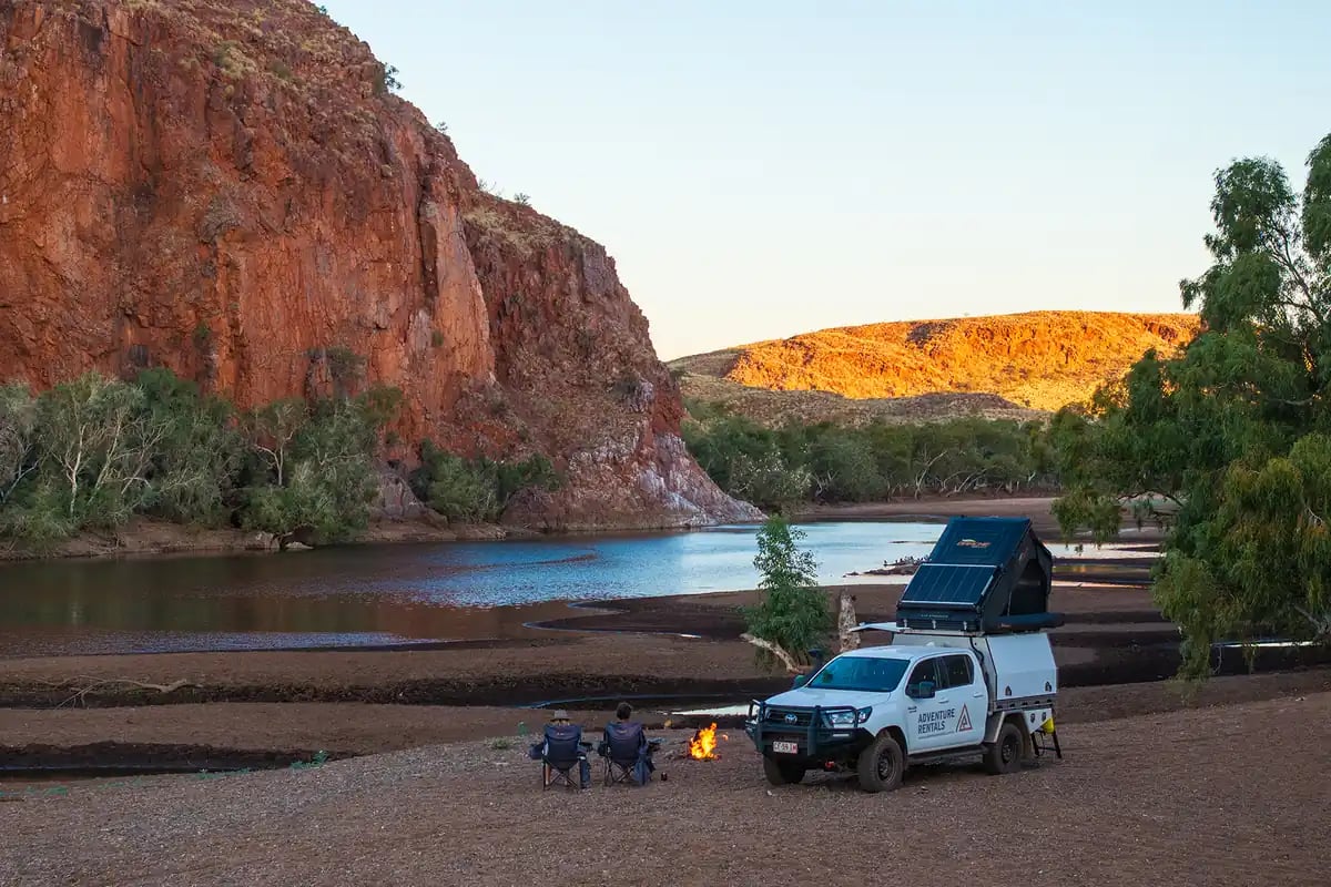 a serene outdoor scene at a riverside campsite. The foreground features a white SUV with a black awning, parked next to a tent that has a blue awning. There are two people sitting on chairs, enjoying the view.