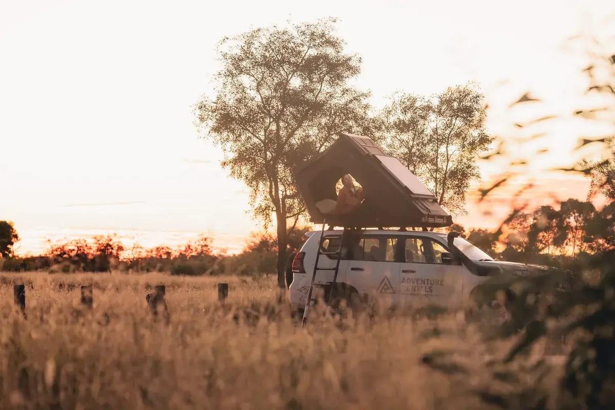 a white adventure camper van parked in a field with tall grass and trees in the background. The van's roof is open, inviting the viewer into the scene. The sky is painted with the warm hues of either sunrise or sunset, suggesting the beginning or end of a journey.
