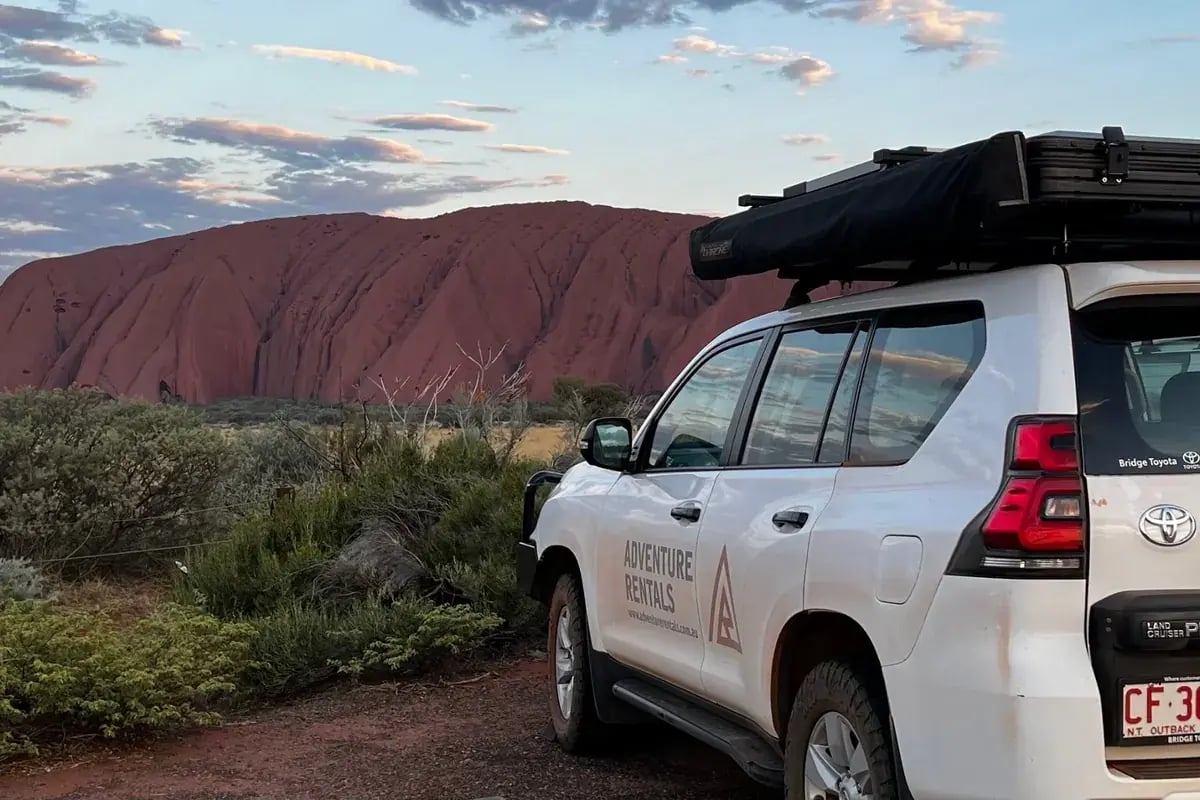 a white SUV with 'ADVENTURE TRAILS' written on its side, parked in a natural setting with red rock formations in the background, indicating a landscape that could be associated with outdoor or adventure activities.