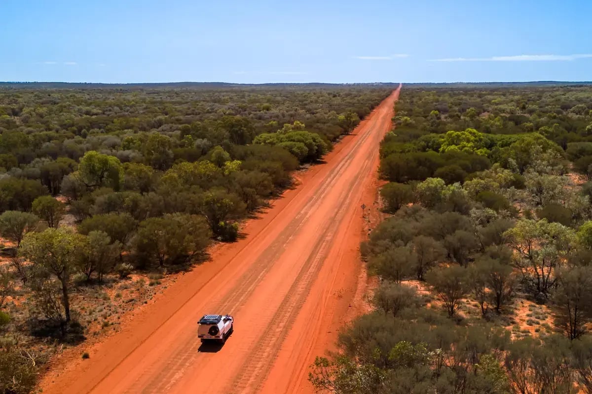 an expansive view of a long, straight dirt road that cuts through a vast desert landscape. A vehicle is driving towards the camera, and the area is dotted with green shrubbery. The sky is clear with no clouds, and the road extends into the horizon, suggesting an open and remote environment.