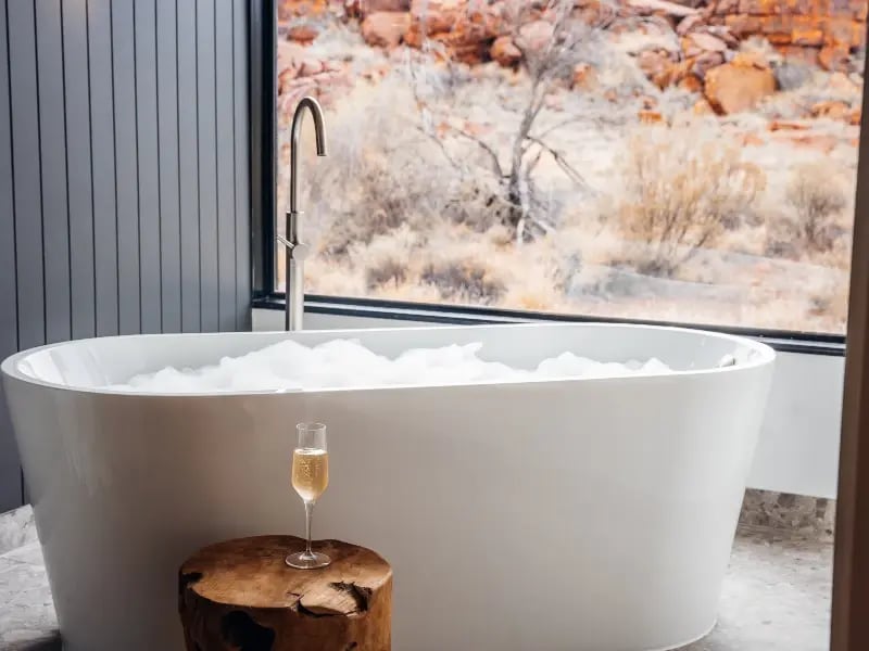 A modern bathroom with a large white bathtub filled with bubbles, a glass of champagne on a wooden stool, and a wall mural of a desert landscape.