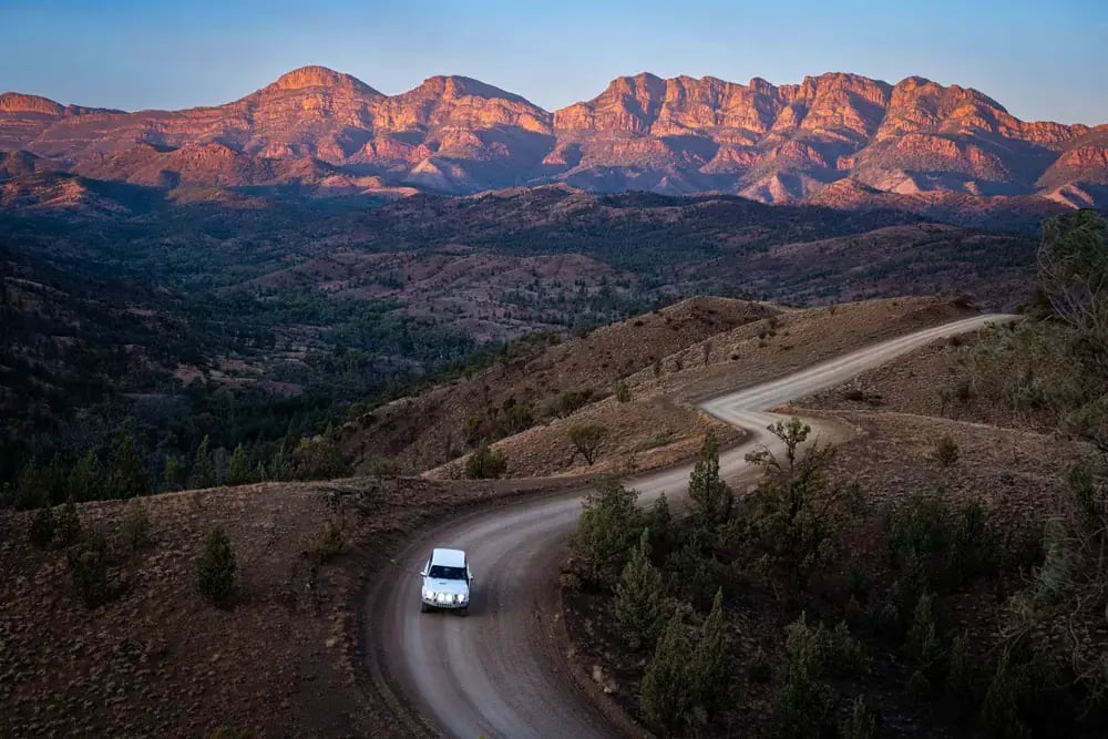 Grusvei med en bil som slynger seg mot fjellkjeden i Flinders Ranges i kveldslys.