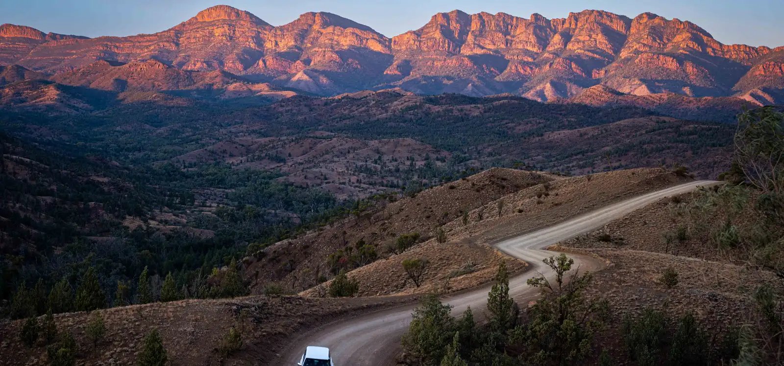 Grusvei med en bil som slynger seg mot fjellkjeden i Flinders Ranges i kveldslys.