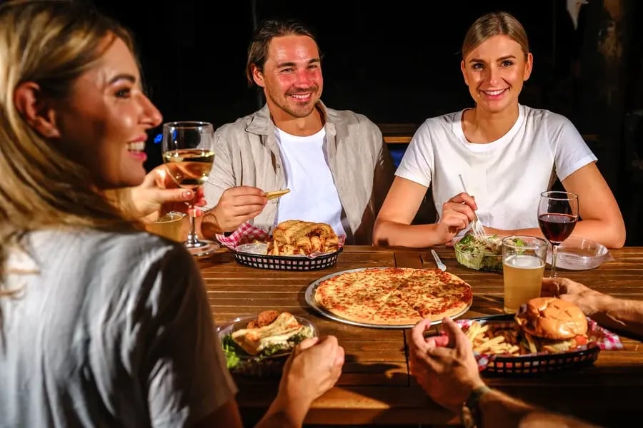 Tre gjester ved et bord med pizza, burgere og drinker.