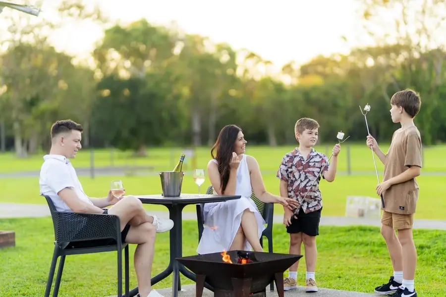 Familie med vin og marshmallows rundt et bålfat på plenen.