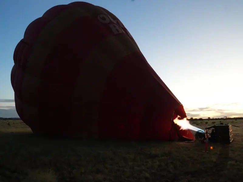 a large, red hot air balloon with the number '8' visible on its side, partially uninflated and lying on the ground. The setting appears to be an open field with a clear sky, possibly during sunrise or sunset due to the warm lighting