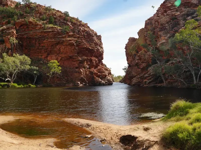 a natural landscape featuring a river with clear, dark water flowing between two rocky cliffs. Vegetation is sparse on the cliffs, and the sky is partly cloudy. There is a small, reddish-brown area on the riverbank that could be a rock or sediment pile.