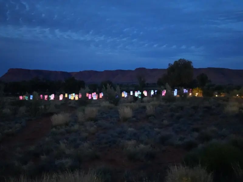 a desert landscape at twilight with a series of brightly lit signs or billboards aligned along a road. The sky is dusky, and the landscape includes arid terrain with sparse vegetation. The lights create a contrast against the darker surroundings, suggesting a rural or less populated area.