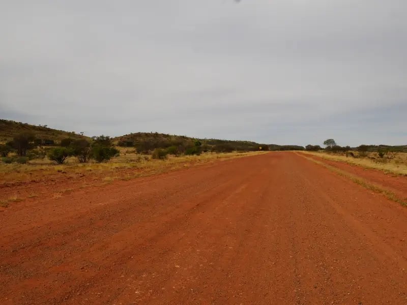 a long, straight dirt road that stretches into the distance. The road has a rough texture and is surrounded by sparse vegetation, primarily small bushes and shrubs. The sky is overcast, suggesting a cloudy day.