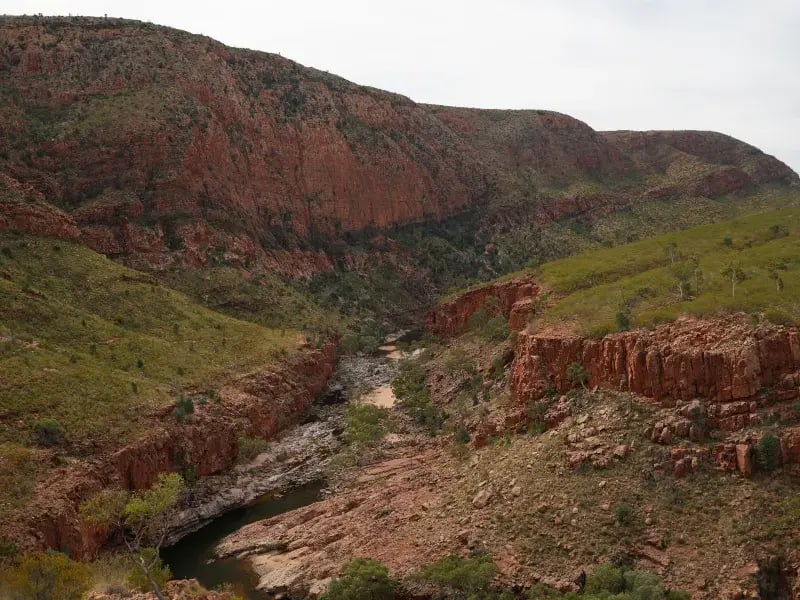 a natural landscape featuring a river cutting through a valley with steep, layered rock formations. The rocks are predominantly in shades of red and brown, and the riverbed is visible with water flowing through it.