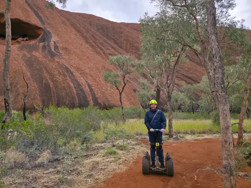 a person engaged in Segway touring, navigating through a red sandy terrain with distinctive red and orange cliffs in the background. The person is equipped with a helmet, indicating safety precautions while on the Segway.