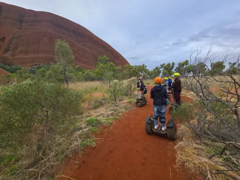 a group of people on hoverboards navigating a red dirt path in a natural, hilly outdoor setting.