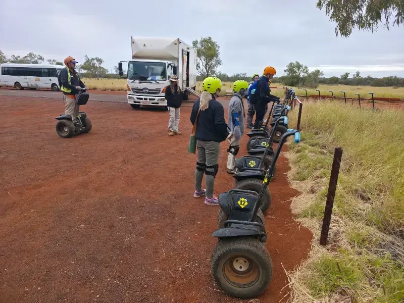 a group of people standing in a line on a dirt path, with a series of black, wheeled devices with yellow tops positioned in front of them - segways.