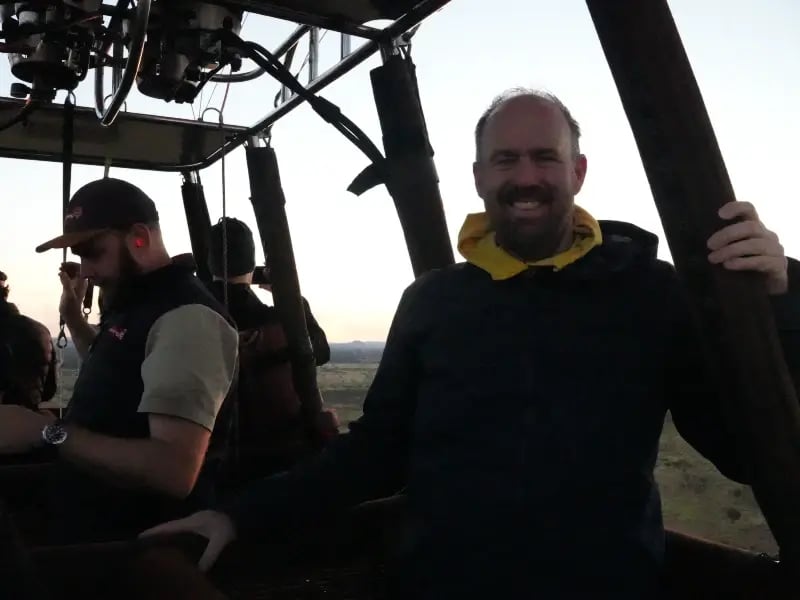 two men inside a hot air balloon, with one man in the foreground smiling and wearing a black jacket, and the other in the background with a cap and a dark jacket.