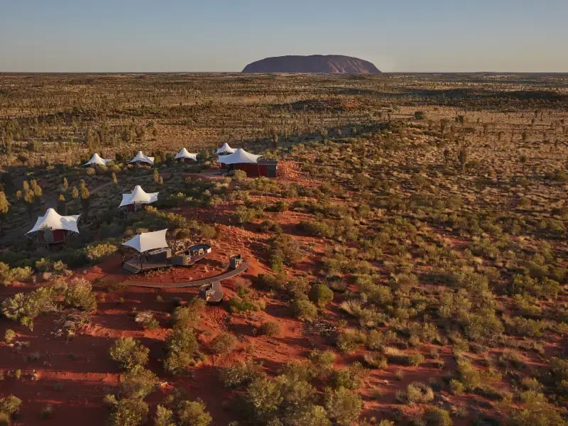 a remote desert landscape with a series of white tents and a red structure, possibly a tented accommodation or a station, set up on a dirt road. In the background, there is a large, rounded rock formation that resembles Uluru. The terrain is arid with sparse vegetation, and the sky is clear.