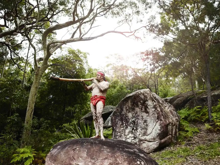 Mann spiller didgeridoo ved steinblokker i skogen.