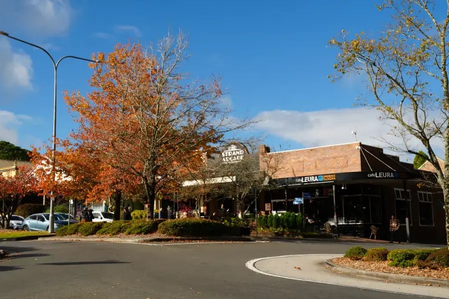 a sunny day at a shopping center with a clear blue sky and a few clouds. There are several trees with autumn-colored leaves, and a roundabout is visible in the foreground.