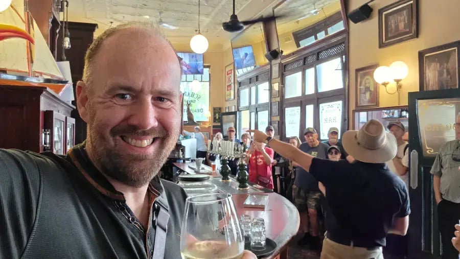 an indoor scene, likely a bar or restaurant, with a person in the foreground wearing a black shirt and holding a glass of white wine.