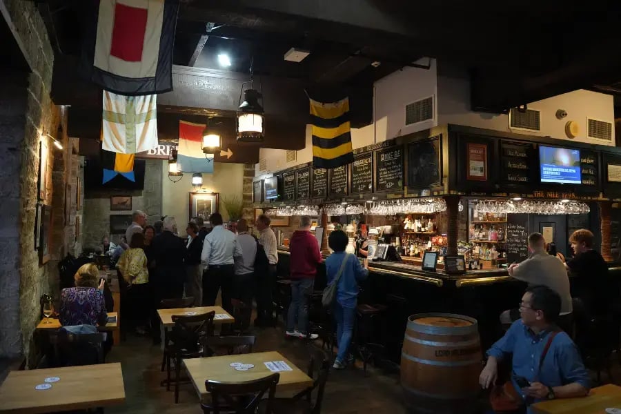 an indoor bar or restaurant scene with patrons gathered around the bar area. There are wooden tables and bar stools, a variety of flags and banners, and a menu board hanging above the bar. A person is seated at the bar, and the overall ambiance suggests a casual dining or socializing environment.